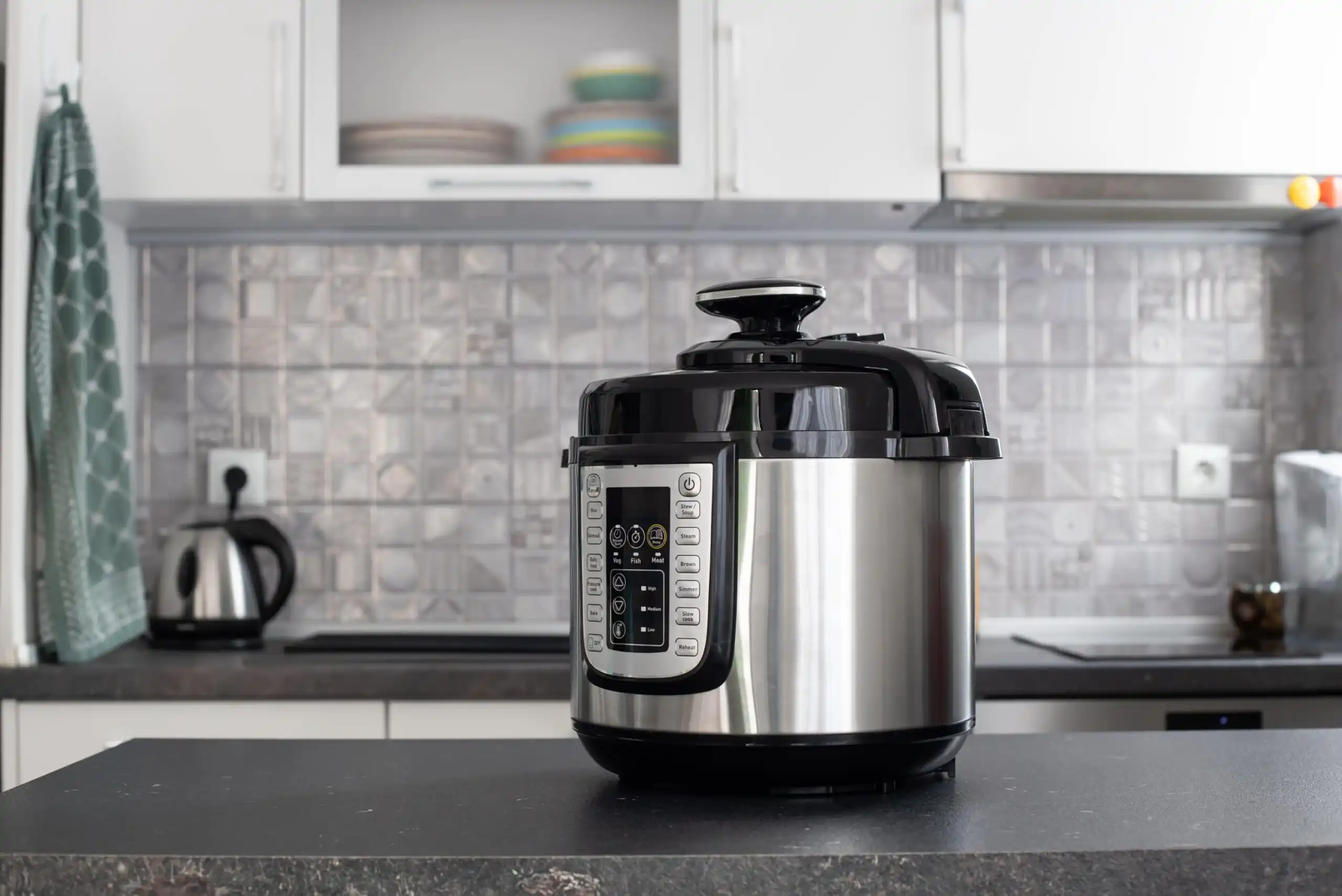 A modern electric pressure cooker with a digital control panel sits on a dark kitchen countertop; in the background are white cabinets, a kettle, and a towel hanging on a hook.