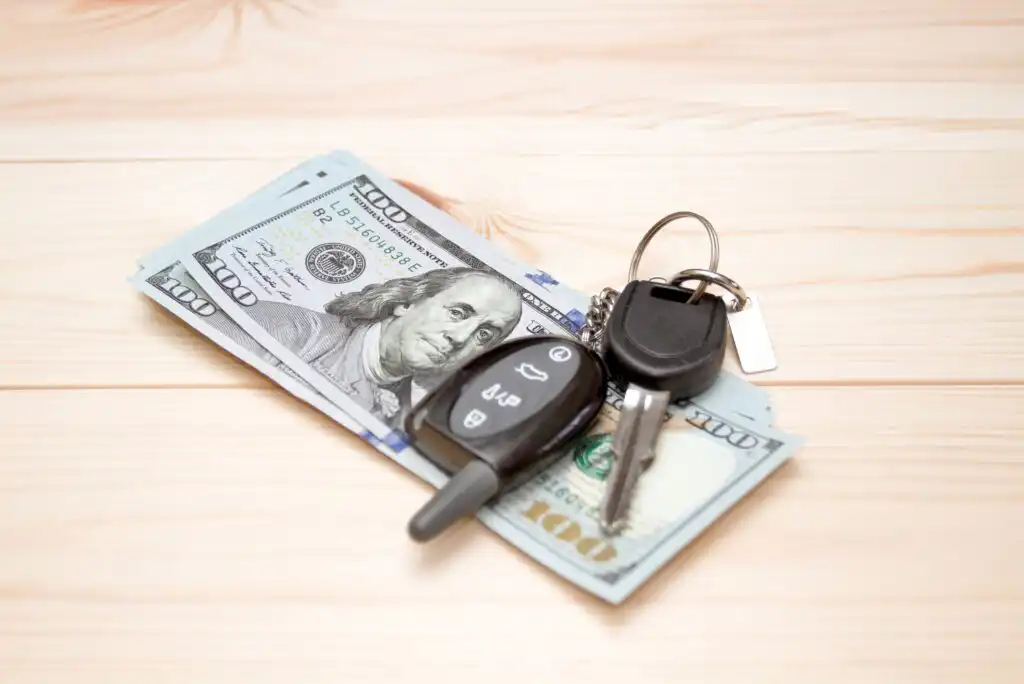 Two car keys and a remote key fob are placed on top of several one-hundred-dollar bills on a light wooden surface.