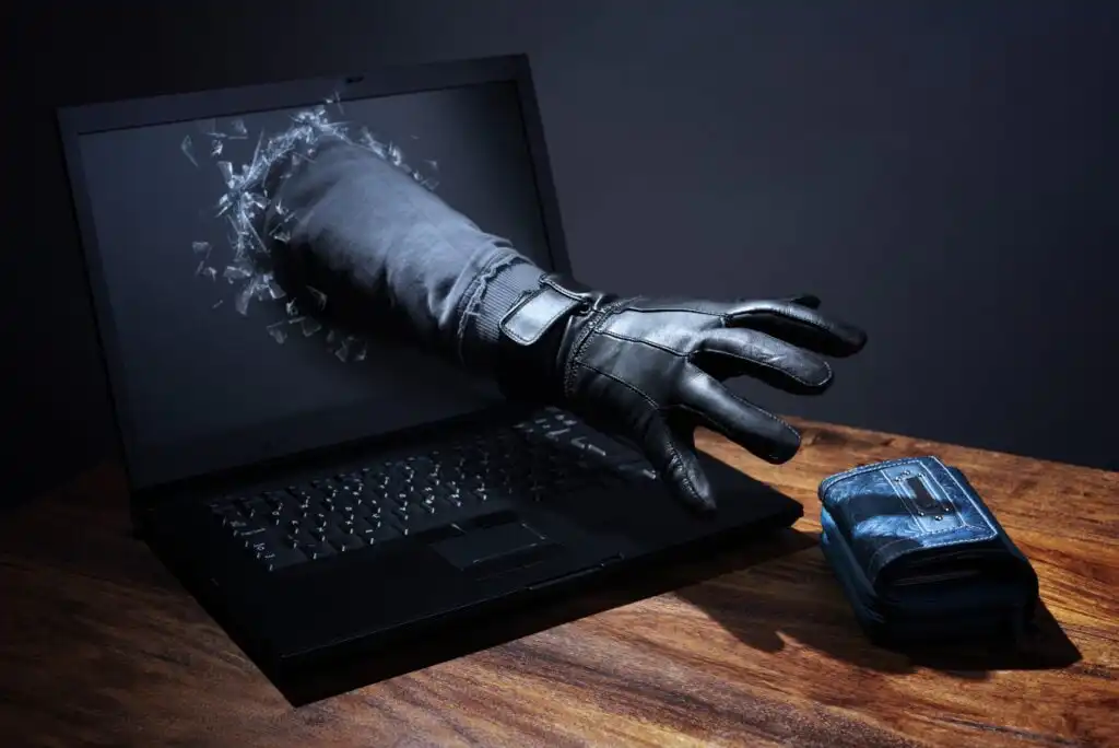 A gloved hand is emerging from a broken laptop screen, reaching toward a wallet on a wooden table, symbolizing cyber theft or hacking.