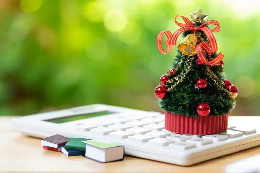 A small decorated Christmas tree sits on a calculator, with miniature books nearby, against a blurred green background.