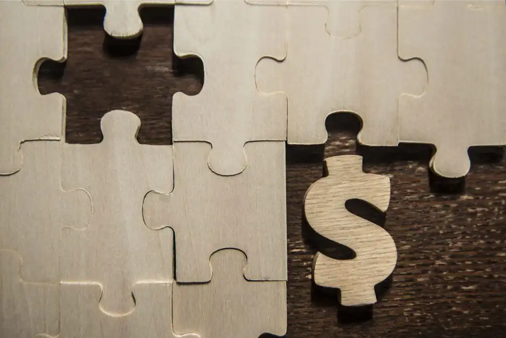 Several beige wooden puzzle pieces are arranged on a dark wood surface, with one puzzle piece shaped like a dollar sign placed among them, symbolizing the concept of financial solutions or investment.