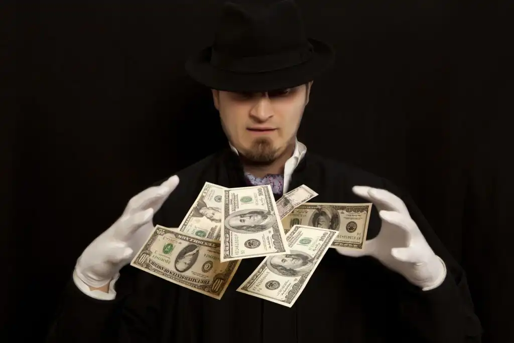 A man in a black hat and coat with white gloves appears to magically levitate several U.S. dollar bills in front of him against a dark background.