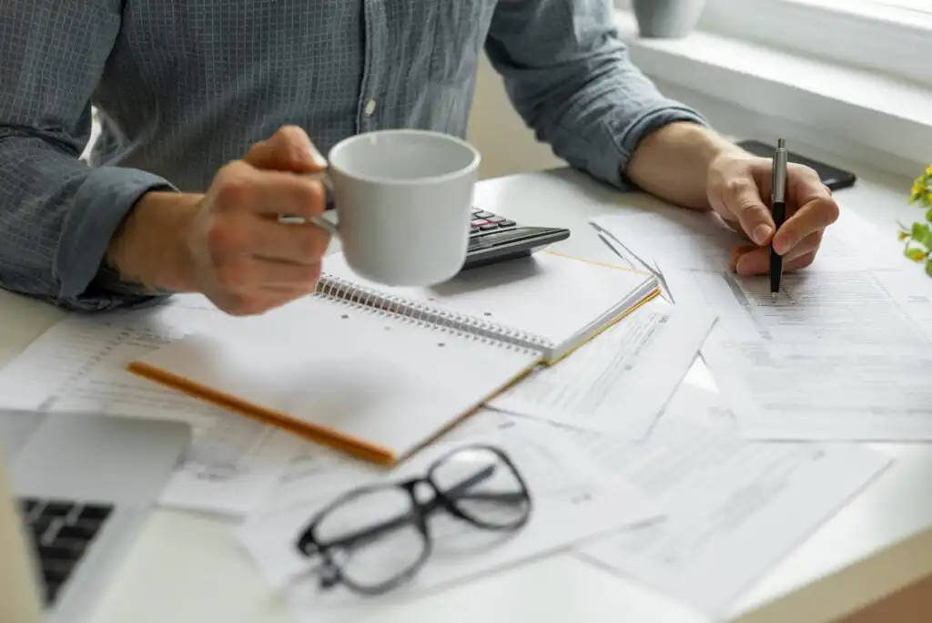A person holds a white coffee mug while reviewing documents and writing on paper at a cluttered desk with notebooks, a calculator, and eyeglasses.