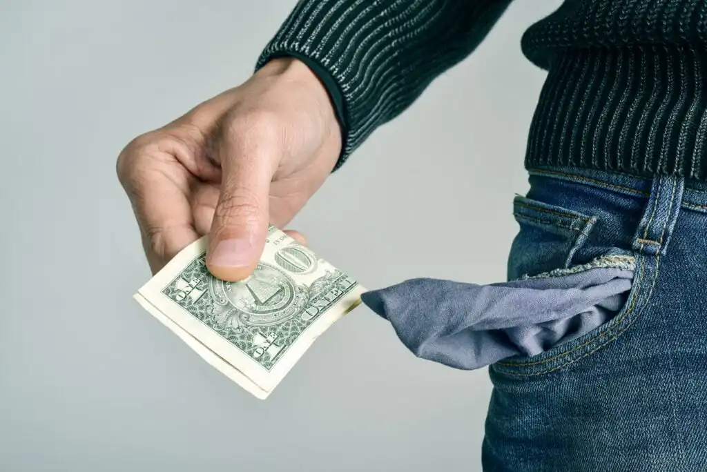 A person wearing jeans with an empty, turned-out pocket holds a single US dollar bill, suggesting financial struggle or having little money.