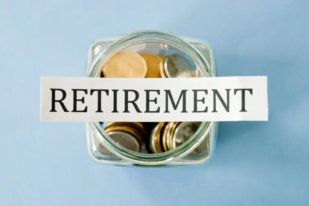 A glass jar filled with coins is labeled with a strip of paper that reads RETIREMENT on a light blue background.