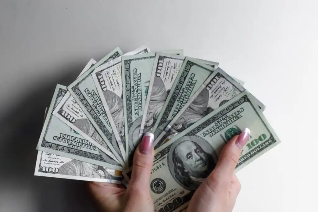 A person with manicured nails holds a fanned-out stack of U.S. one hundred dollar bills against a plain white background.