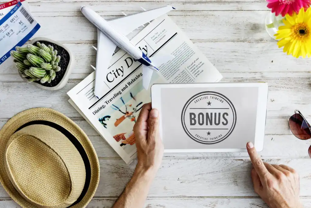 A person holding a paper with the word BONUS beside a straw hat, sunglasses, a pen, a plant, a boarding pass, a newspaper, and yellow flowers on a white wooden table.