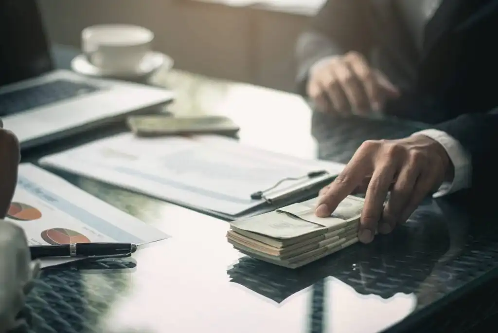 A person’s hand reaches for a stack of cash on a desk with documents, a pen, and a laptop, suggesting a business transaction or financial deal. Another person sits across the table, partially visible.