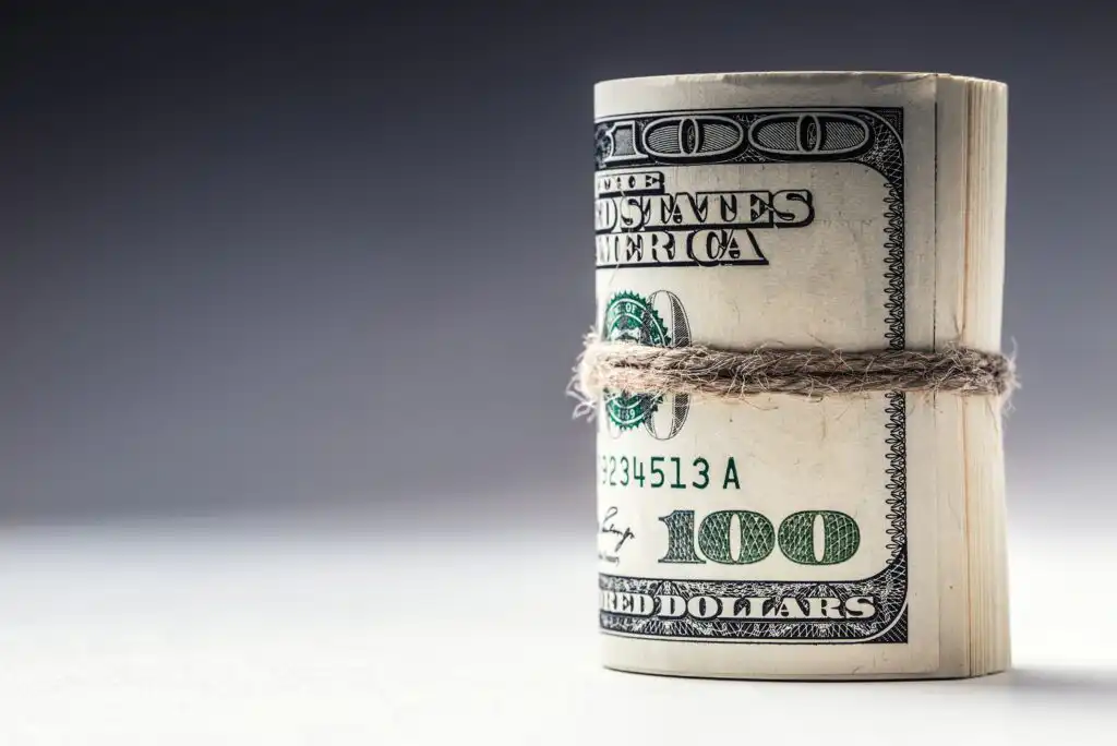 A roll of 100 US dollar bills wrapped with a piece of twine, standing upright on a plain surface against a dark, gradient background.
