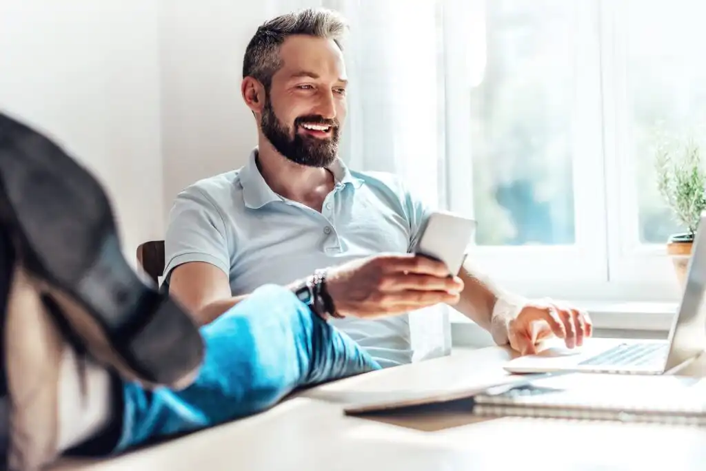 A smiling man with a beard sits at a desk with his feet up, holding a smartphone in one hand and using a laptop with the other. Sunlight streams through a window, giving the room a bright, relaxed feel.
