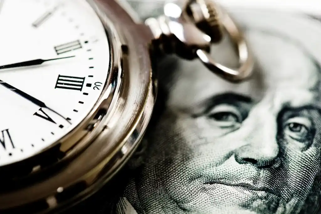 Close-up of a pocket watch showing Roman numerals next to a portion of a U.S. hundred dollar bill featuring Benjamin Franklin’s face, symbolizing the concept of time and money.