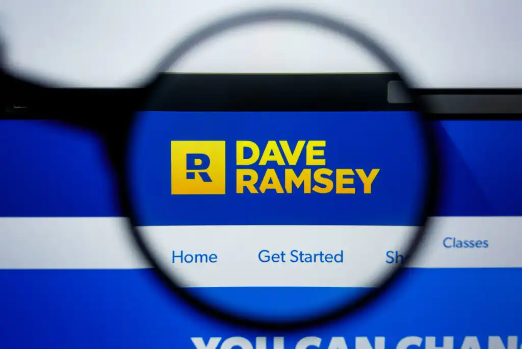 A magnifying glass focuses on the “Dave Ramsey” website logo in yellow and blue, showing navigation options: Home, Get Started, and Shop, against a blurred website background.