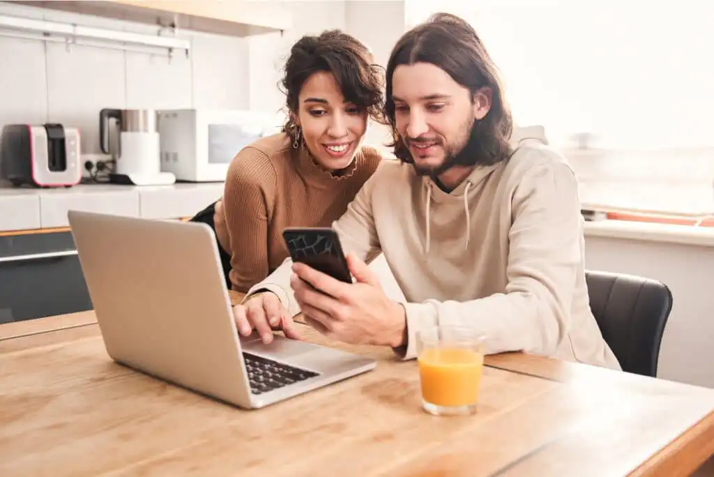 A smiling couple sits at a kitchen table, looking at a smartphone together. A laptop is open in front of them and a glass of orange juice is on the table. The kitchen is bright and modern.
