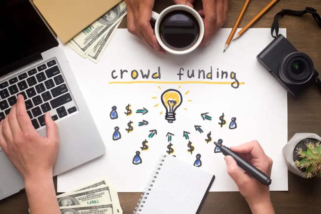 A desk with a laptop, camera, cash, coffee, and a notepad, featuring a paper titled “crowd funding” with drawings of people, dollar signs, and arrows pointing to a light bulb, symbolizing idea funding.