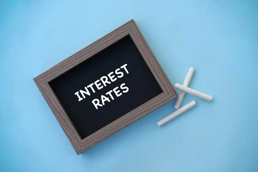 A small chalkboard with the words INTEREST RATES written on it, lying on a light blue surface next to three pieces of white chalk.