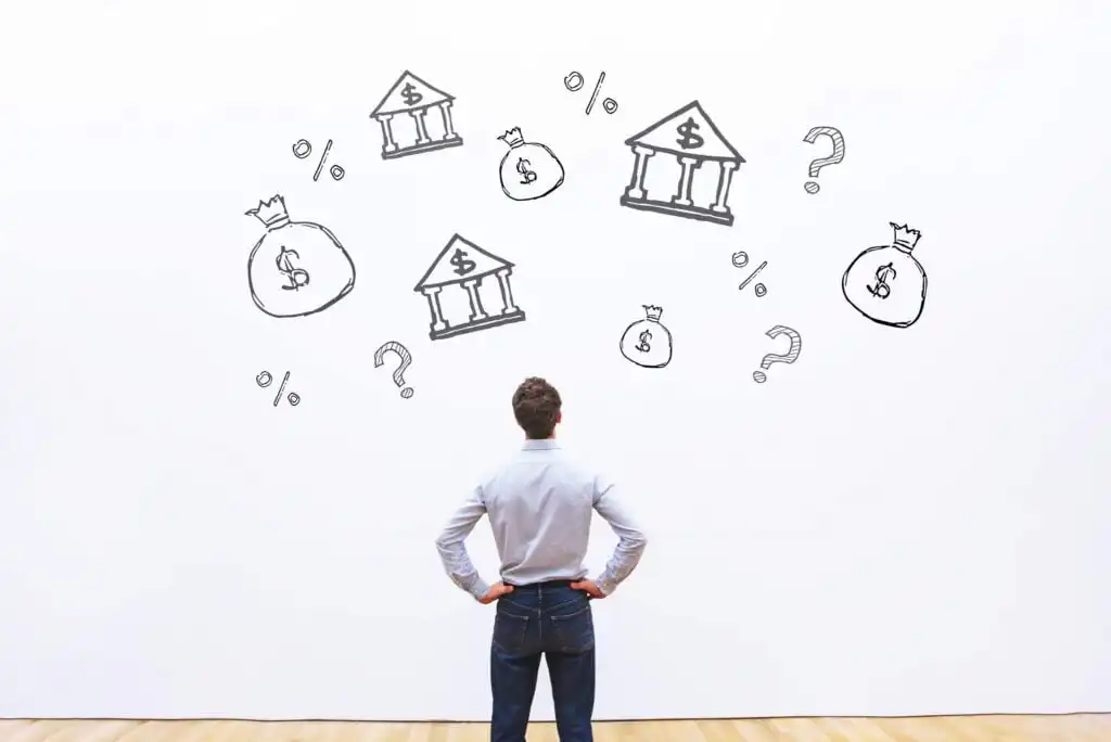 A man stands with hands on hips, looking at a white wall filled with sketches of banks, money bags, dollar signs, percentage symbols, and question marks, representing financial decision-making or planning.