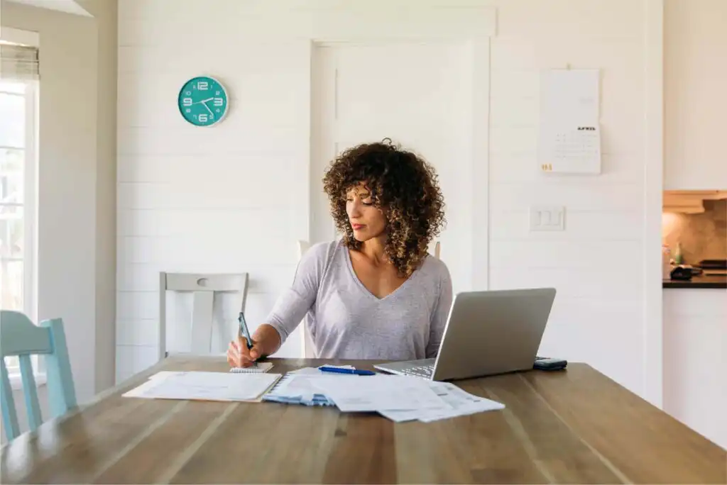 A woman sits at a wooden table working with papers and a laptop. She writes in a notebook, with bills spread out, in a bright room with a clock on the wall and a calendar in the background.