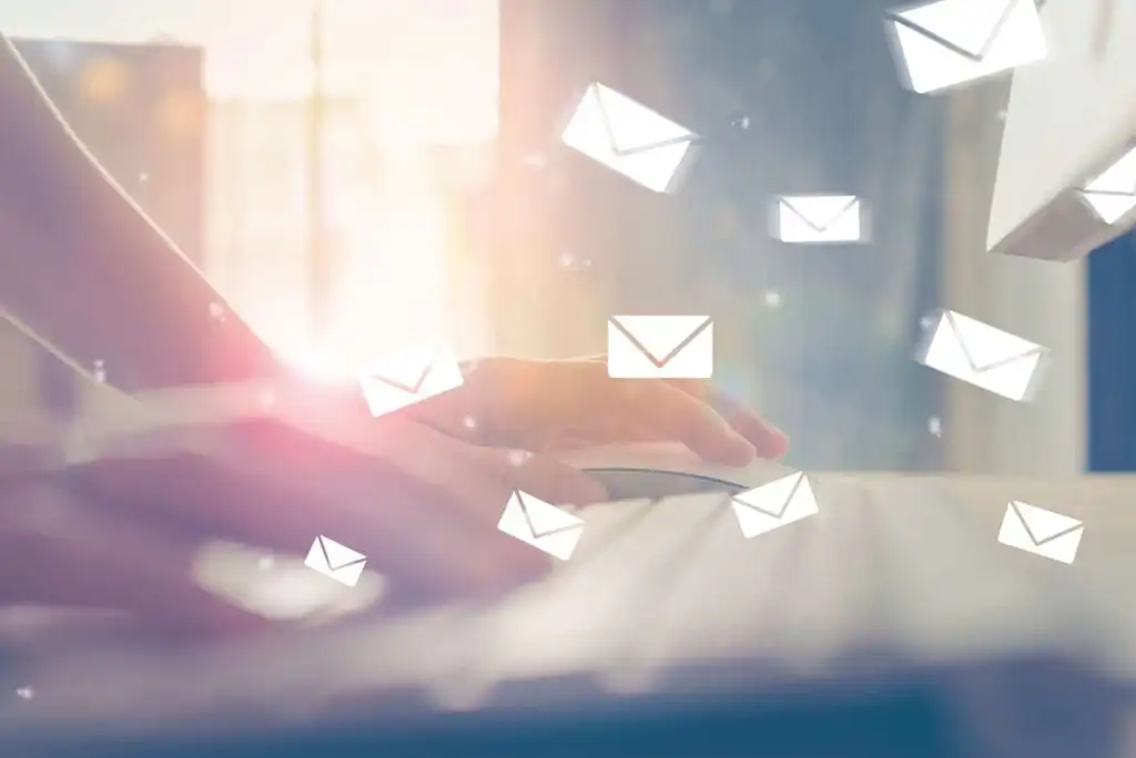 A person’s hand uses a computer mouse beside a keyboard, with illustrated email icons floating in the air, symbolizing sending or receiving emails in a bright, sunlit office setting.