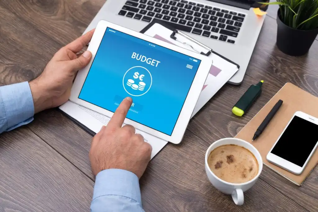 A person uses a tablet displaying a budget app at a wooden desk with a laptop, clipboard, pen, highlighter, and a cup of coffee. The app screen has a piggy bank icon and dollar symbols.