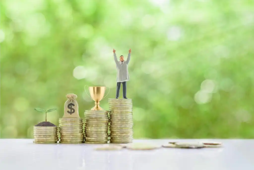 A small figure of a person stands on top of stacked coins beside a trophy, a money bag, and a coin stack with a sprouting plant, symbolizing financial growth and success, with a green blurred background.