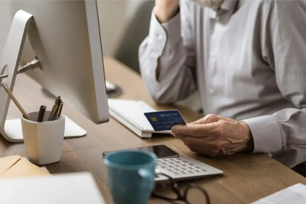 A person sits at a desk with a computer, holding a credit card in one hand and typing with the other. The desk has a calculator, notepad, mugs, and office supplies. The setting looks like a home or office workspace.