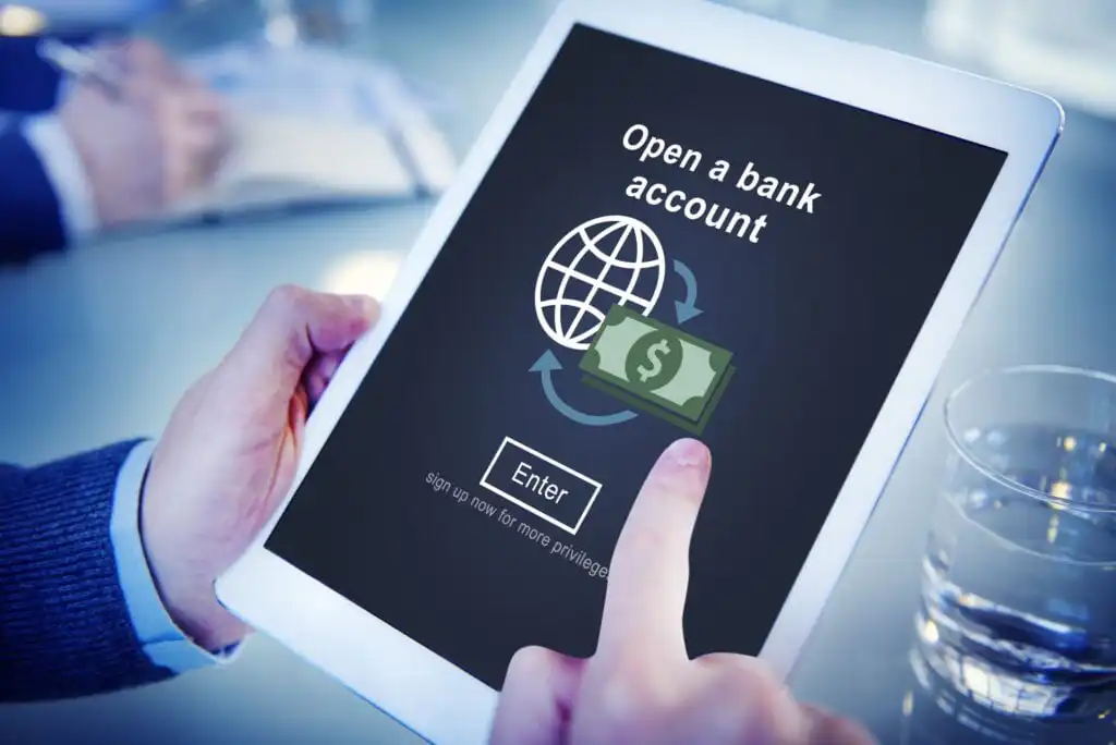 A person holds a tablet displaying a webpage to open a bank account, featuring a globe, dollar bill icon, and an Enter button. A glass of water and another person’s hands are visible in the background.