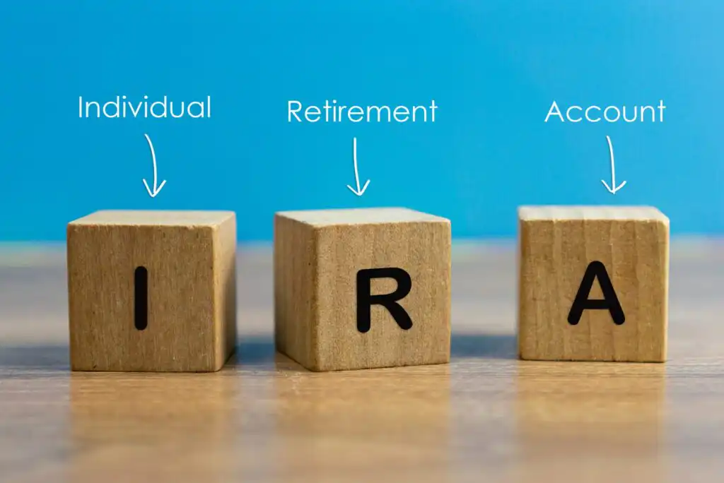 Three wooden blocks labeled I, R, and A sit side by side on a wooden surface. Above each block are the words “Individual,” “Retirement,” and “Account” with arrows pointing to each letter. Blue background.