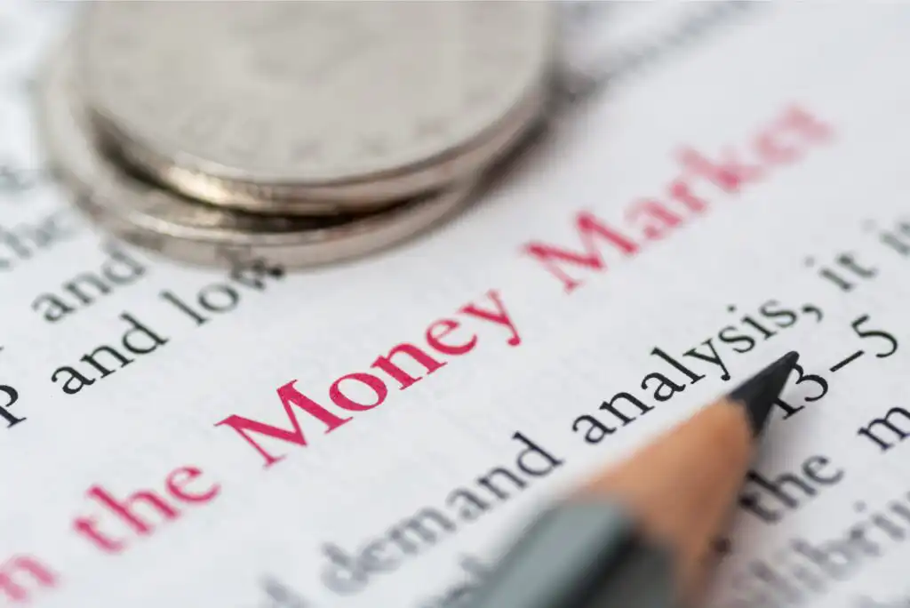 Close-up of a pencil pointing to the words Money Market highlighted in red text on a printed page, with blurred coins stacked in the background.