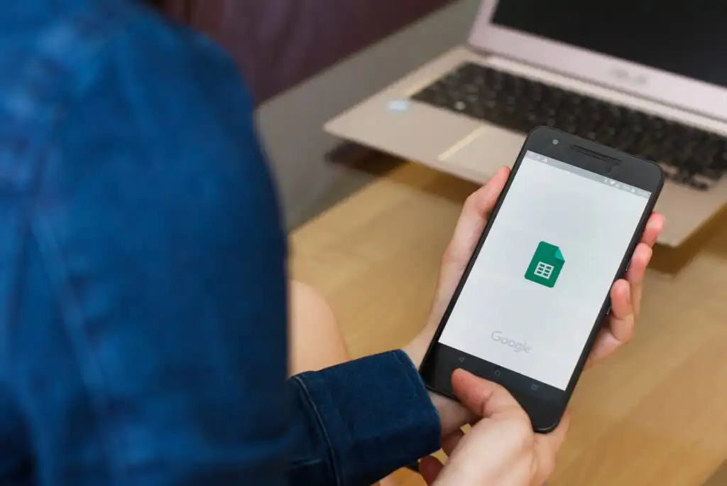 A person holding a smartphone displaying the Google Sheets app logo, with an open laptop on a wooden table in the background.