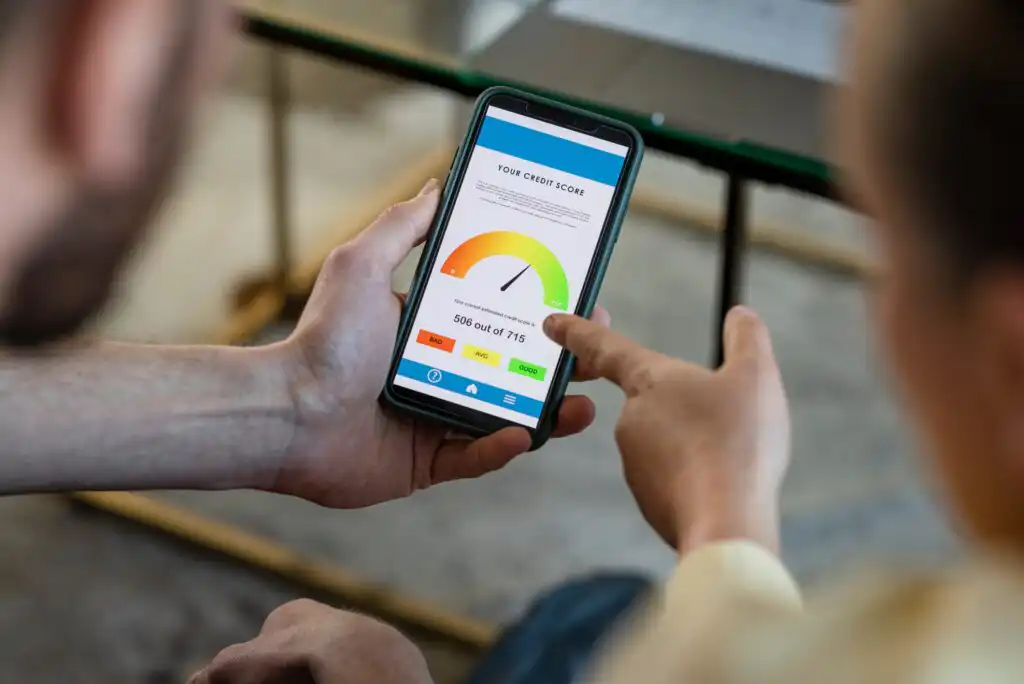 A person holds a smartphone displaying a credit score screen, with a colorful gauge showing a score of 540 out of 715. Another person points at the phone. The setting appears informal, possibly an office or home.