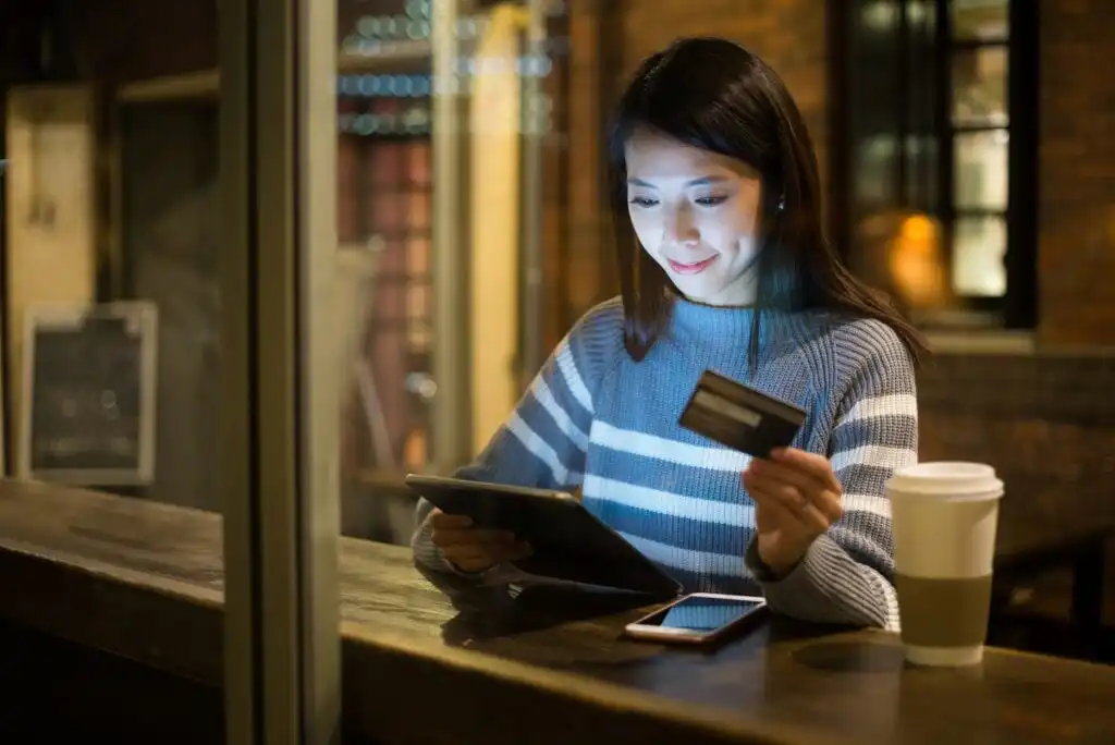 A woman sits at a table by a window, holding a credit card and using a tablet. A smartphone and a takeaway coffee cup are on the table, and she is smiling at the screen in a cozy, dimly lit setting.