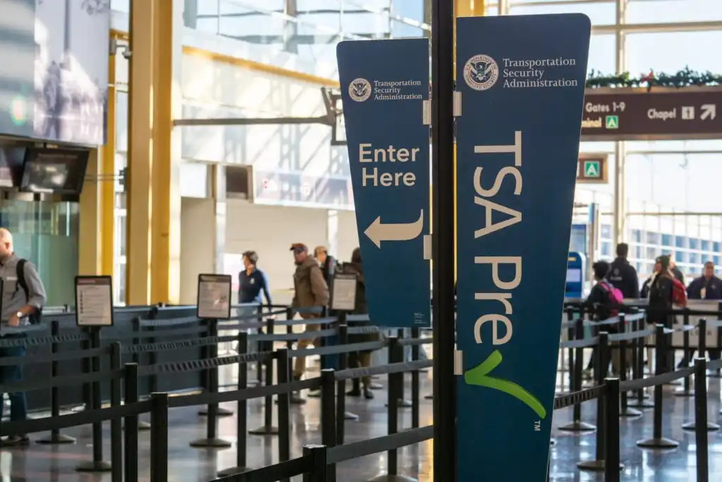 TSA PreCheck and Enter Here signs are displayed at an airport security checkpoint, with passengers waiting in line and a few walking in the background.