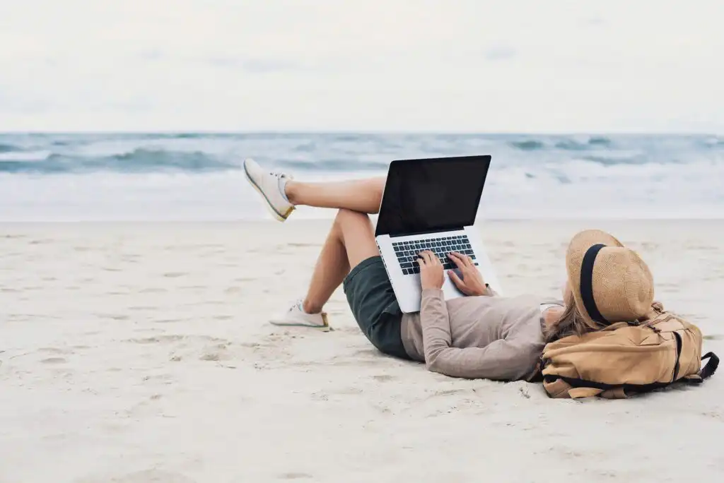 A person lies on a sandy beach near the ocean, using a laptop. They wear a hat, casual clothes, and sneakers, with a backpack beside them. The scene is relaxed and peaceful.