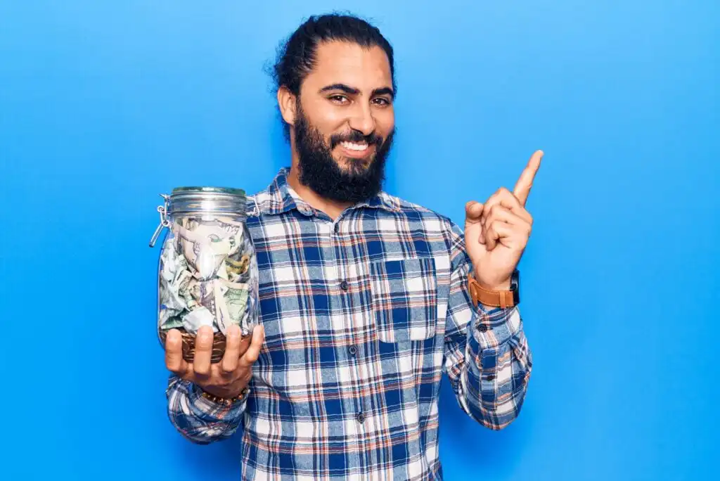 A person with a beard and long hair tied back, wearing a plaid shirt, smiles while holding a jar full of cash in one hand and pointing upward with the other, standing against a bright blue background.