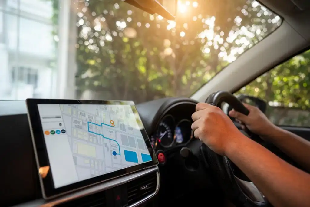 A person driving a car with both hands on the steering wheel, while a GPS navigation system is displayed on a large touchscreen showing a map and route. Sunlight filters through trees outside the window.