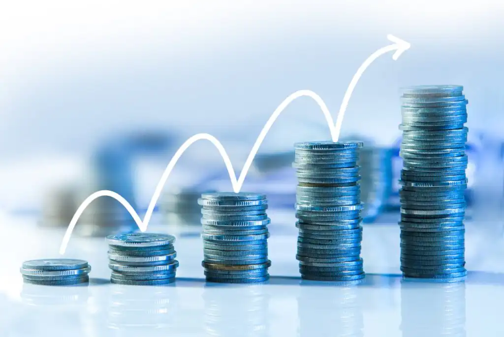 Stacks of coins arranged in increasing height from left to right, with a white upward arrow line illustrating growth or rising profits over time. The background is softly blurred in blue tones.