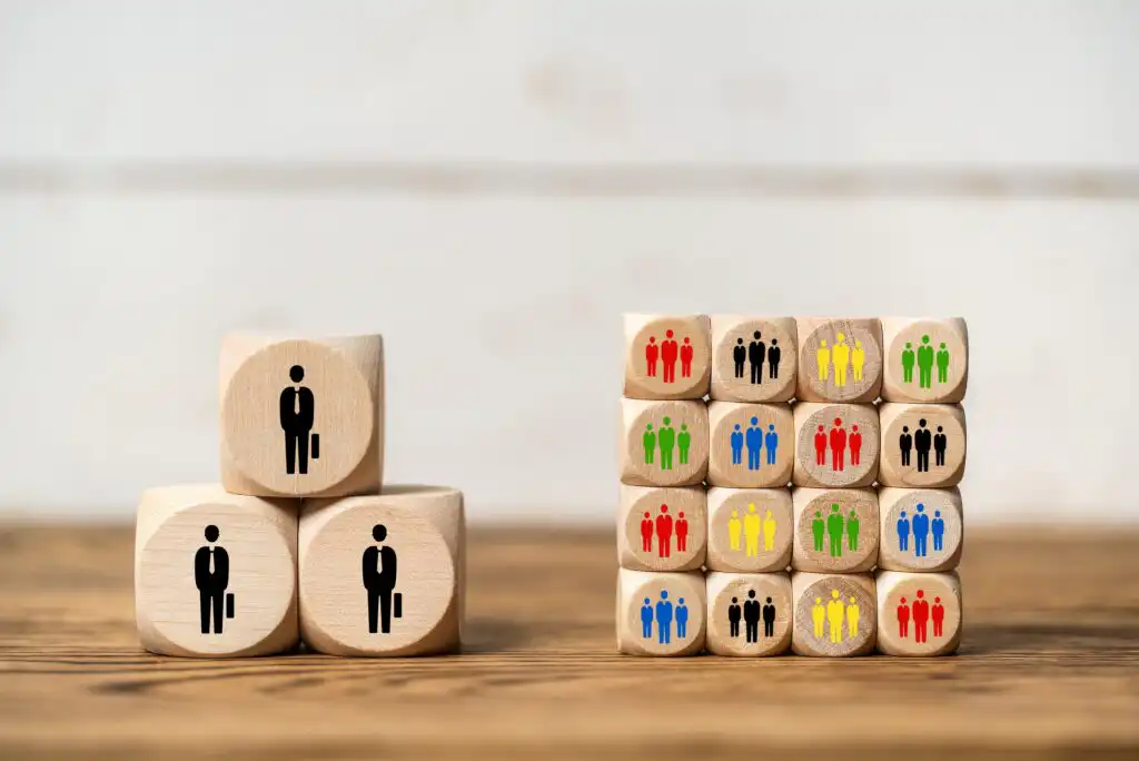 Two wooden blocks with single black stick figures are stacked on the left, while sixteen blocks with groups of colorful stick figures are stacked on the right, representing diversity and teamwork.