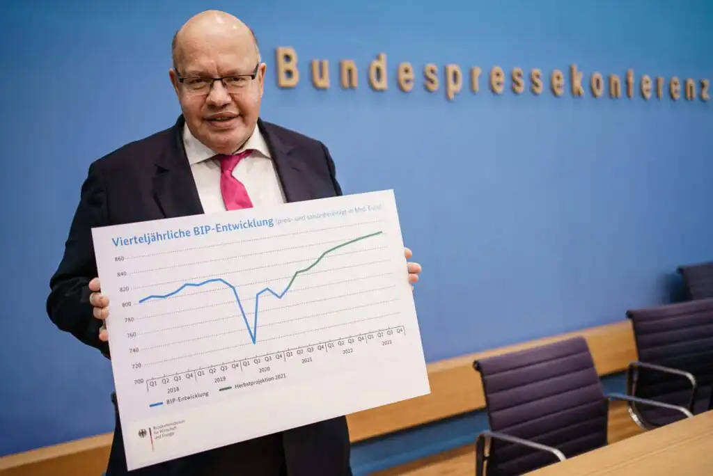A man in a suit and red tie holds a chart showing quarterly GDP development with a sharp dip followed by a rise. He stands in front of a blue wall with the word Bundespressekonferenz in gold letters.