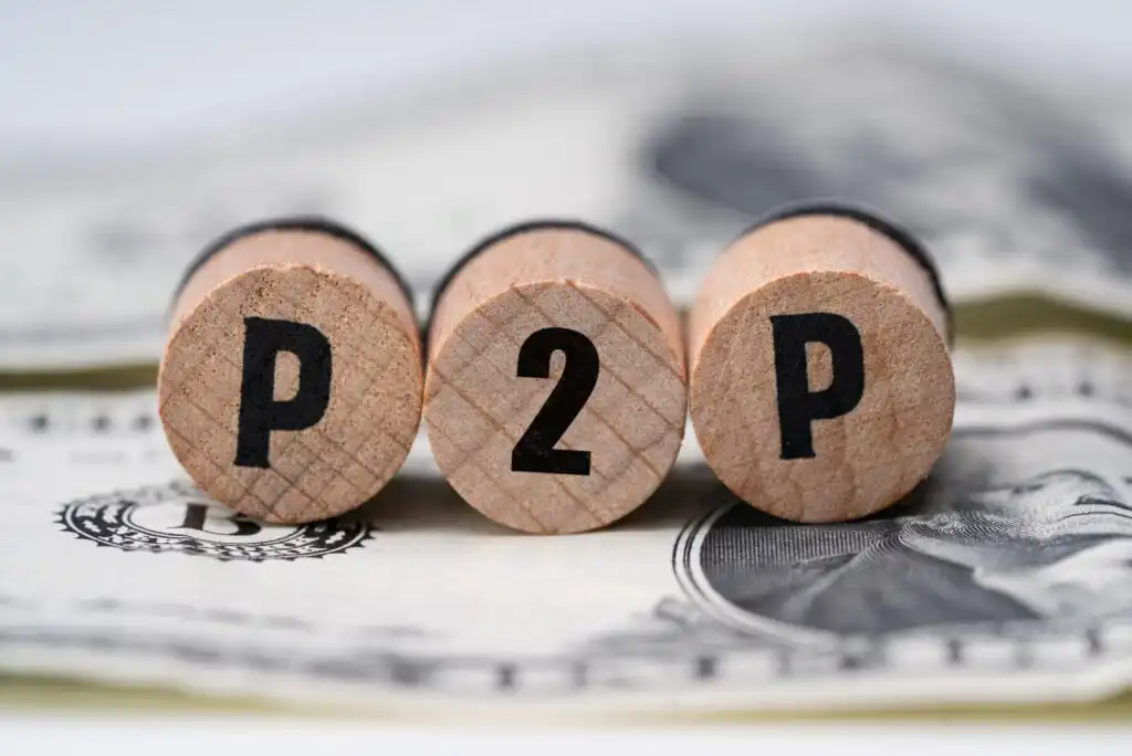 Three wooden blocks with the letters P, 2, and P are arranged in a row on top of a US dollar bill, symbolizing peer-to-peer financial transactions.