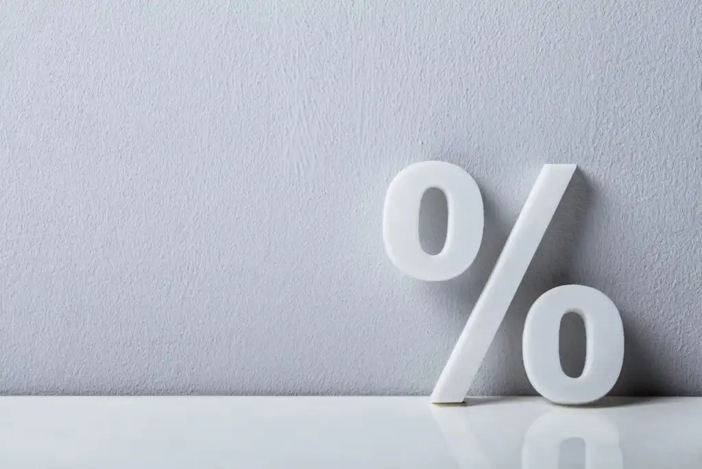 A large white percent symbol (%) stands upright against a plain light gray textured wall, with a reflective white surface below.