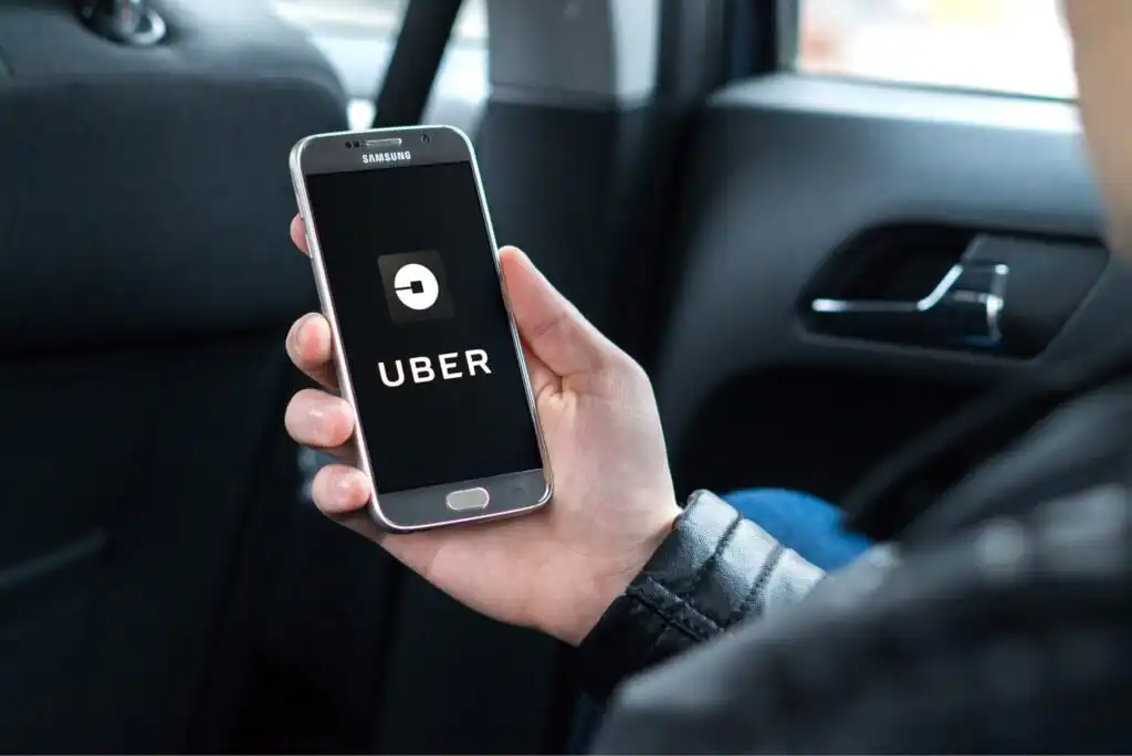 A person sitting in a car holds a smartphone displaying the Uber app logo on the screen.
