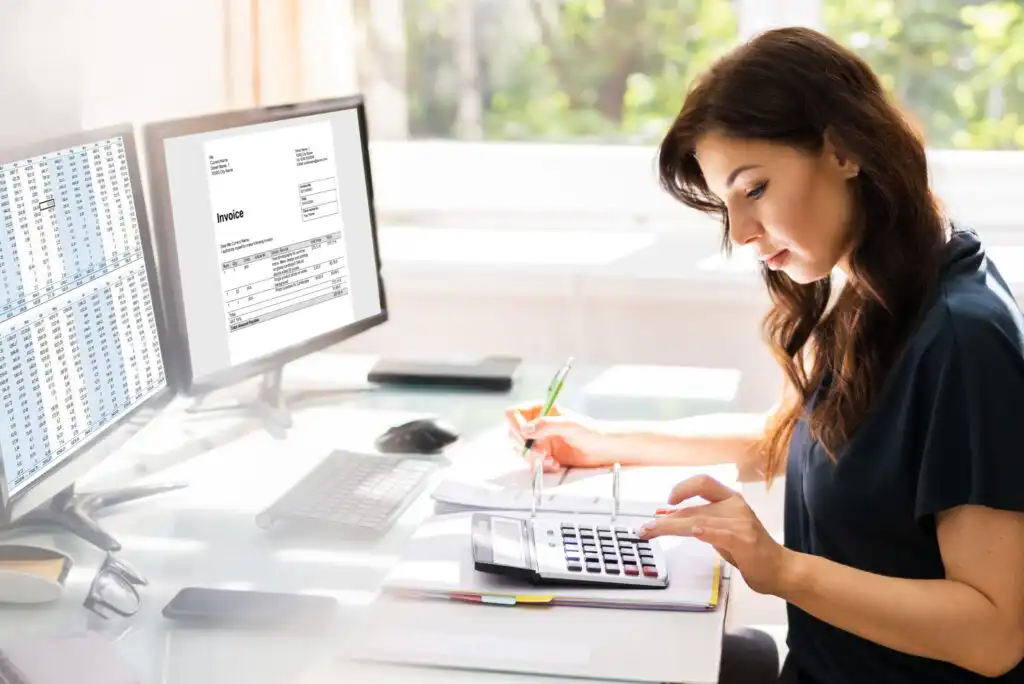 A woman sits at a desk using a calculator, with financial documents and a pen in hand. Two computer monitors display spreadsheets and an invoice. Daylight streams through a window in the background.