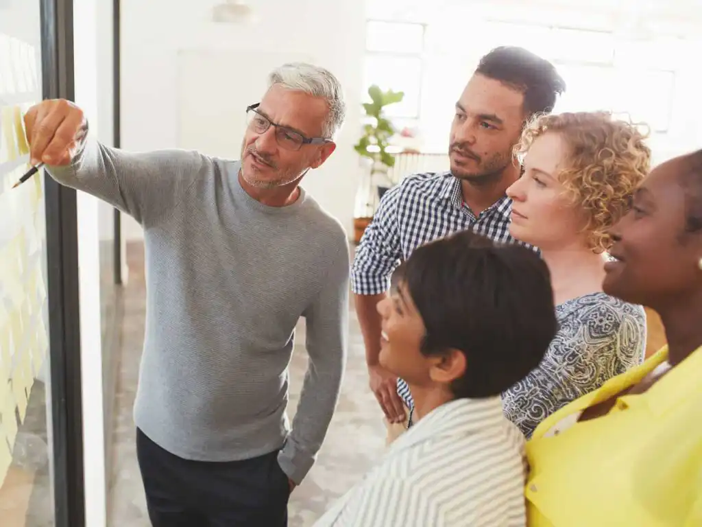A group of five people, diverse in age and ethnicity, stand together looking at sticky notes on a glass wall as one person points and explains an idea during a brainstorming session in a bright office.