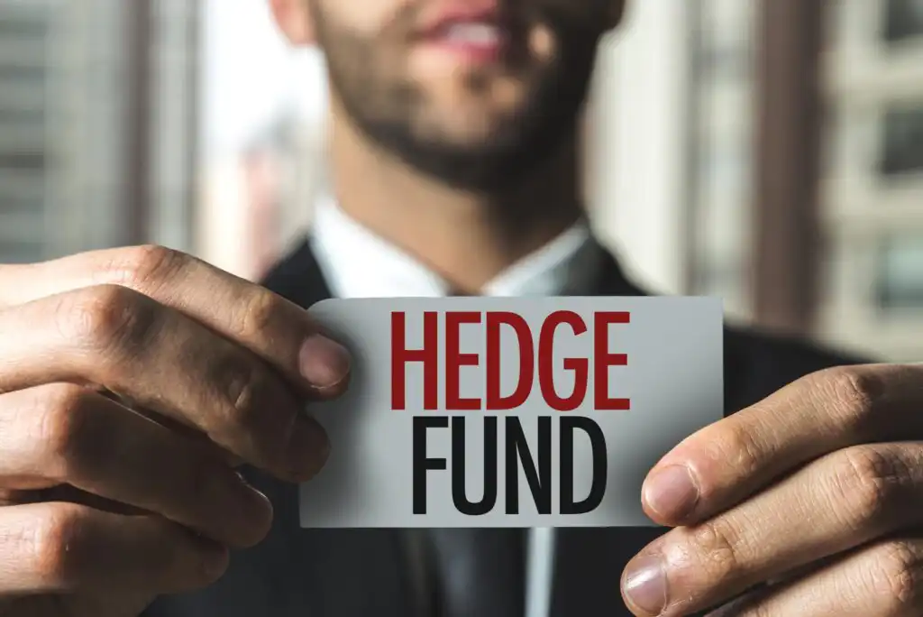 A man in a suit holds a card with the words HEDGE FUND written in bold red and black letters, with a blurred office background.