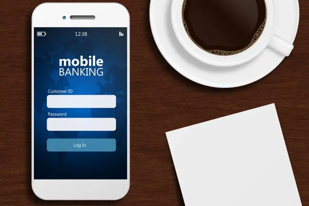 A smartphone displaying a mobile banking login screen sits on a wooden desk next to a cup of coffee and a blank sheet of paper.