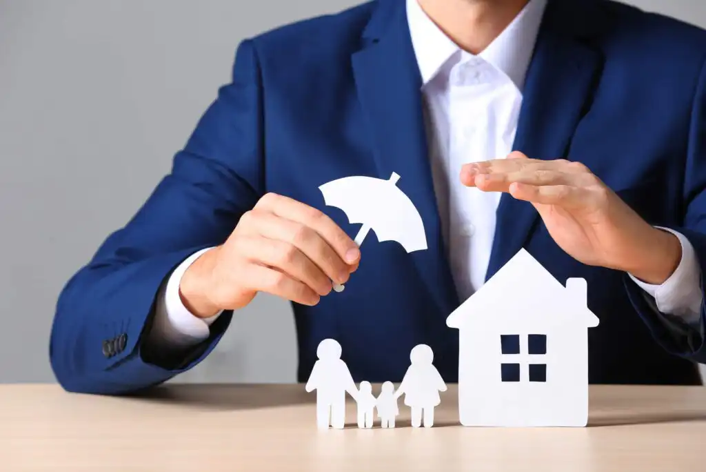 A man in a blue suit holds a paper umbrella over cutouts of a family and a house, symbolizing protection and insurance.