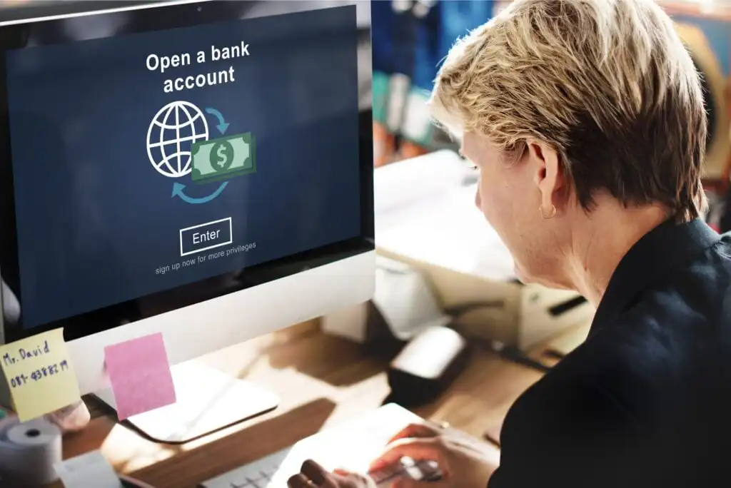 A person with short blonde hair sits at a desk using a computer displaying a screen that says “Open a bank account” with an enter button and bank-related icons.
