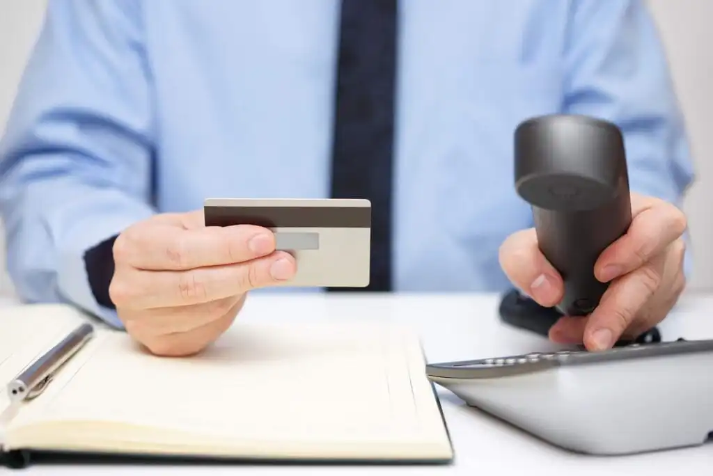A person in a blue shirt holds a credit card in one hand and a payment terminal in the other, sitting at a desk with an open notebook and a pen.