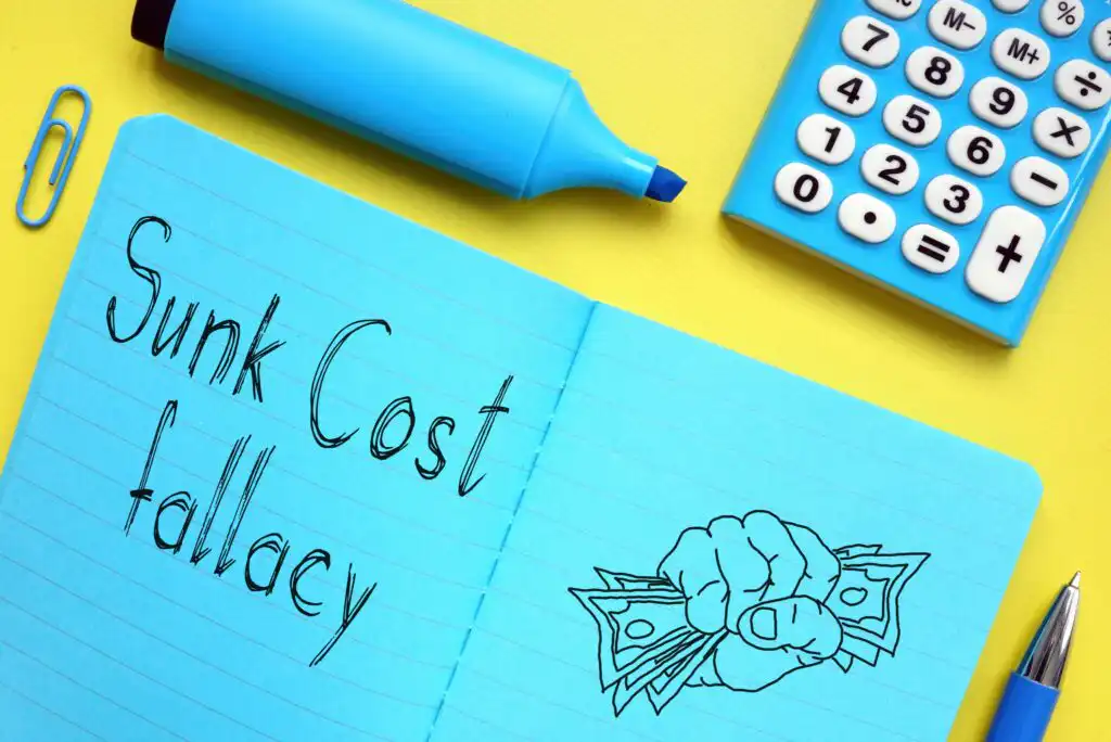 A blue notebook open to a page with Sunk Cost fallacy written and a drawing of a fist holding money, surrounded by a blue highlighter, paperclip, calculator, and pen on a yellow surface.