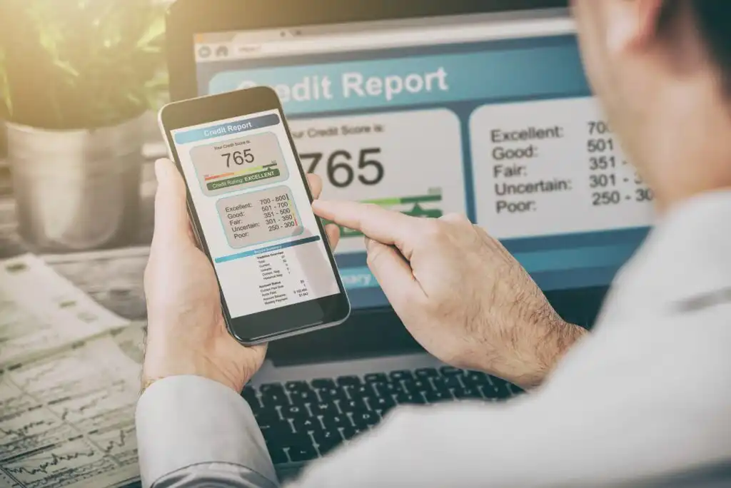 A person checks their credit report on a smartphone while also viewing it on a laptop. The credit score displayed is 765, labeled as excellent, with the screen showing credit score ranges and financial documents nearby.
